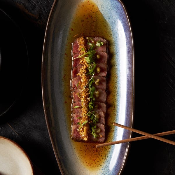 Sliced medium-rare steak topped with crispy onions, scallions, herbs, and green peas on a rectangular ceramic plate with chopsticks.