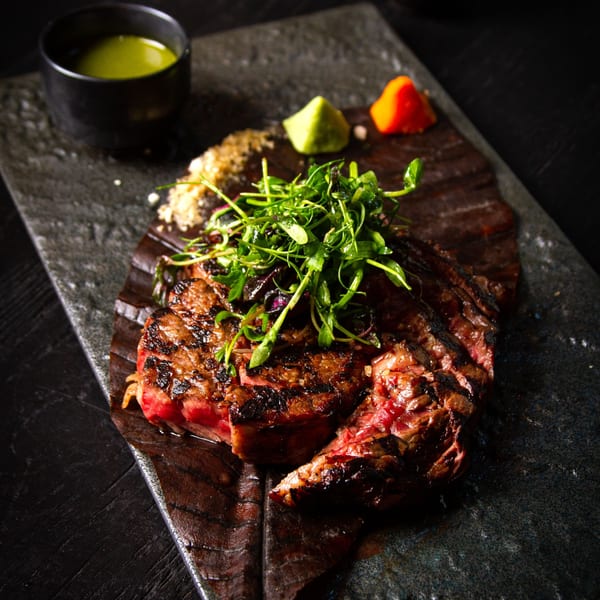 Grilled steak with fresh greens, a side of green sauce, and two colorful condiments on a dark stone plate.