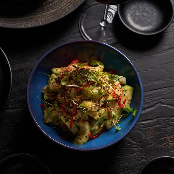 Blue bowl of cucumber salad topped with sesame seeds, sliced red chili, and fresh herbs on a dark wooden table.