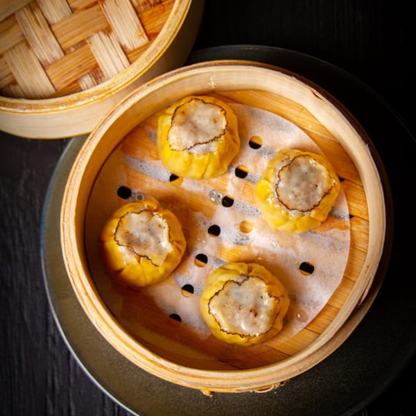 Four steamed dim sum dumplings with yellow wrappers and meat filling in a bamboo steamer.