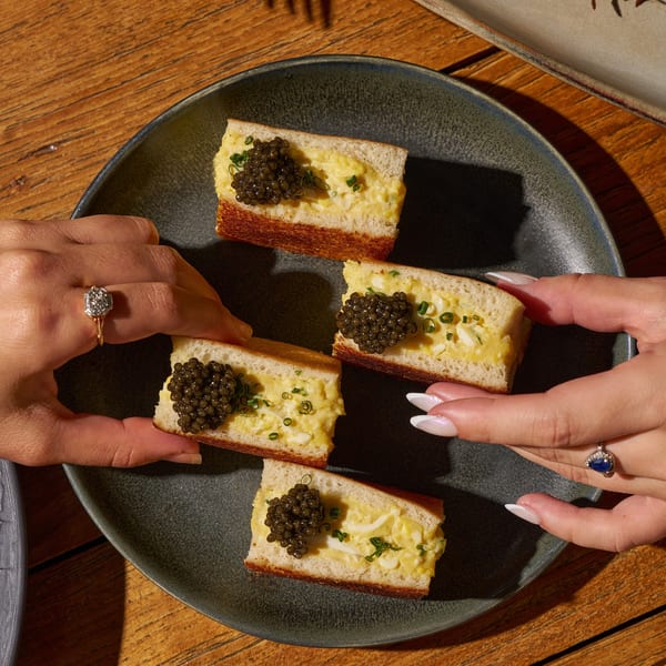 Two people's hands reaching for four pieces of toast topped with scrambled eggs and caviar.