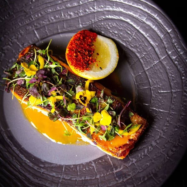 Tanuki River Landing's grilled fish with microgreens, edible flowers, and a lemon wedge on a dark plate.