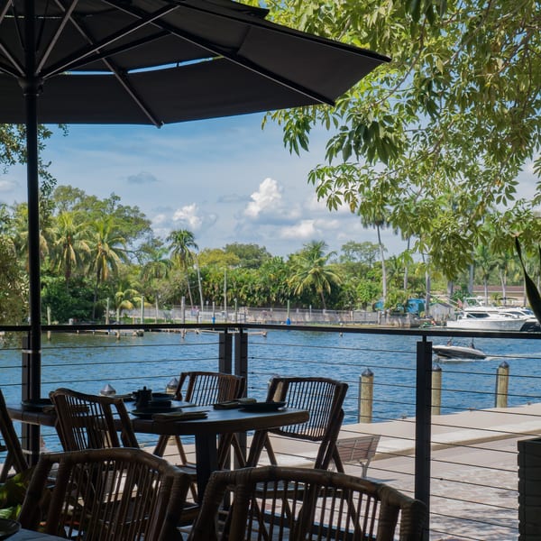 Outdoor dining area with tables and chairs overlooking a river with boats and palm trees.