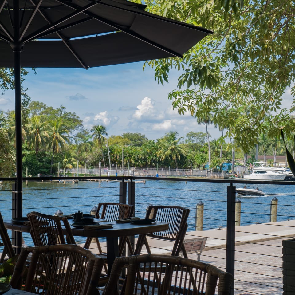 Outdoor dining area with tables and chairs overlooking a river with boats and palm trees.