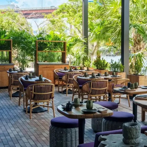 Tanuki River Landing's outdoor dining area with rattan chairs, wooden tables, and lush greenery by the water.