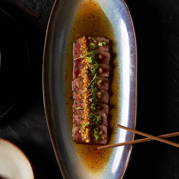 Sliced beef tataki with crispy garlic, scallions, and sauce on a long blue and brown plate.