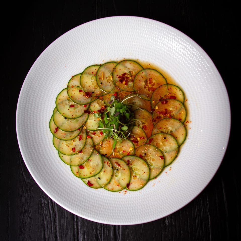 Thinly sliced cucumbers arranged in a circle, topped with chili flakes and microgreens on a white plate.