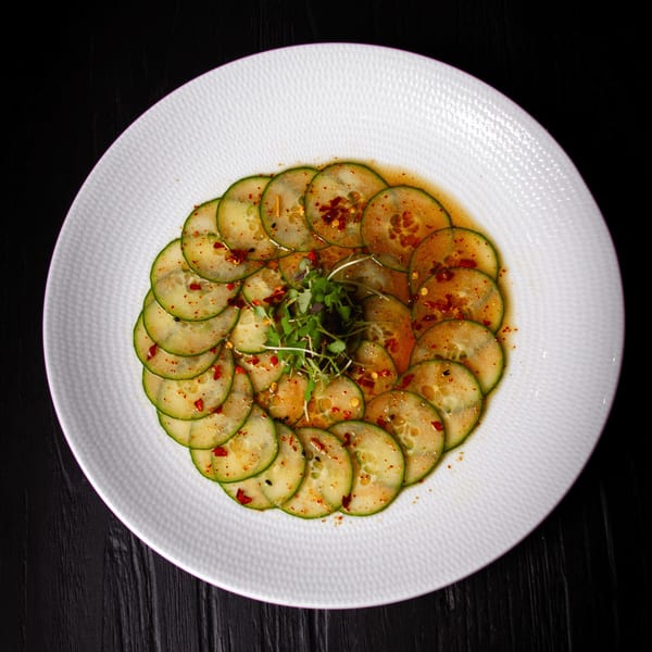 Thinly sliced cucumbers arranged in a circle, topped with chili flakes and microgreens on a white plate.