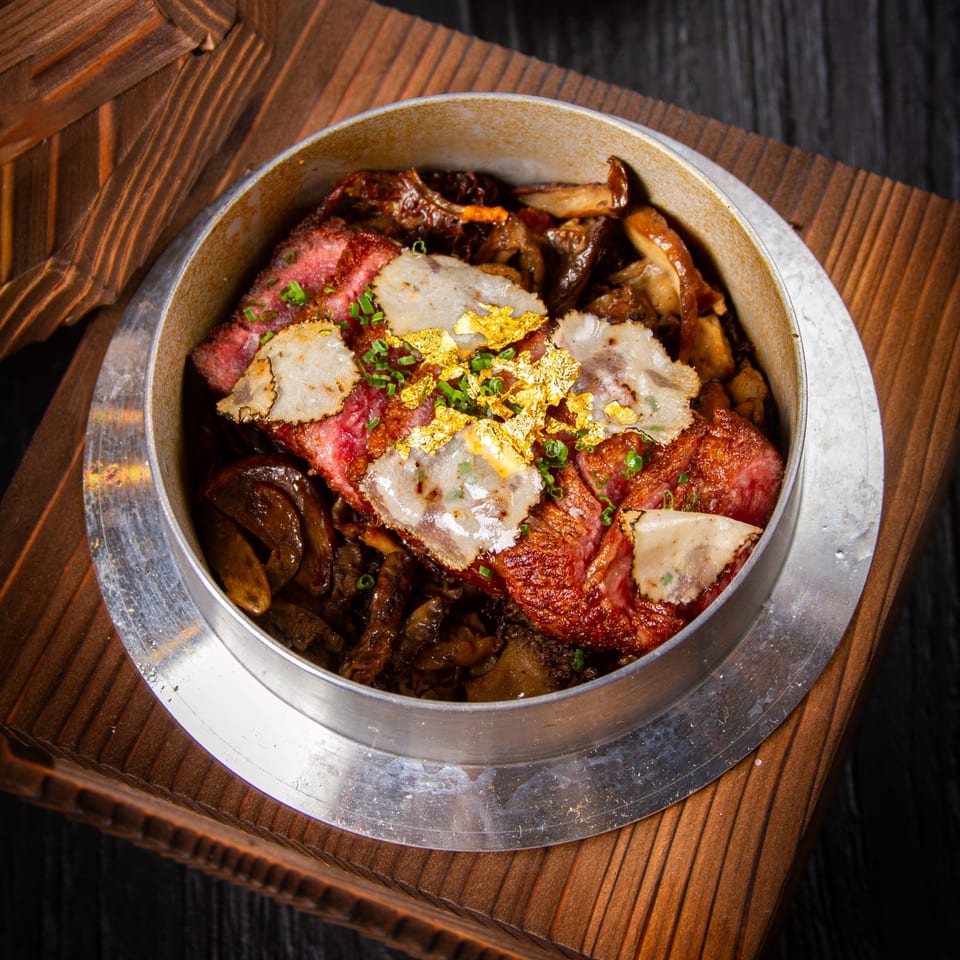 Wagyu beef with mushrooms, truffle slices, and gold flakes in a metal pot on a wooden tray.