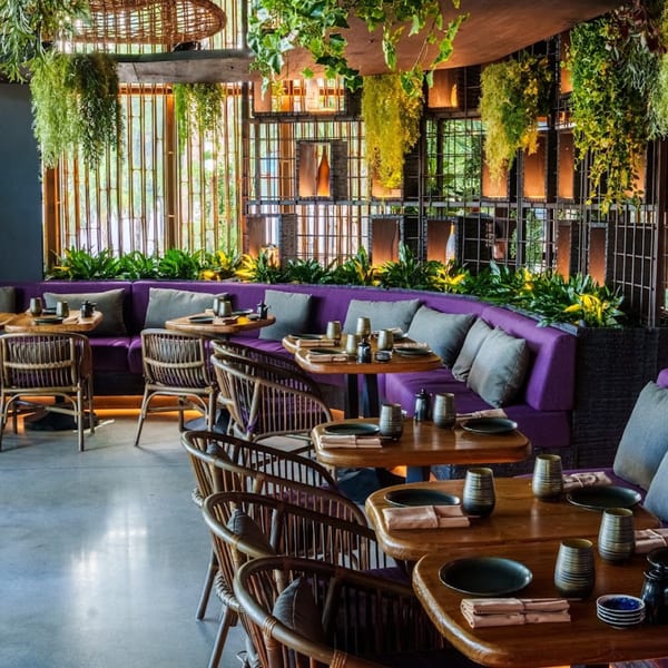 Modern restaurant interior with purple banquettes, wooden tables, rattan chairs, and lush hanging greenery.